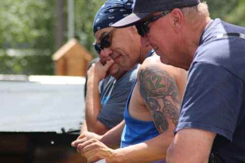 3 men in sunglasses lean on a rail, looking down and smiling. The middle man has a large tribal tattoo on his upper left shoulder.