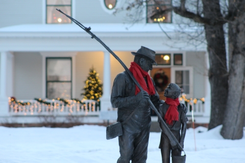A statue of a grandfather taking his granddaughter fishing is spruced up with red scarves tied around their necks. The statue has snow all around it with Christmas lights in the background on a large white house.