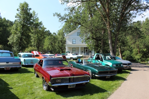 Historic cars and trucks of different makes and colors sit on the green lawn in front of a large white historic house on the hatchery grounds.