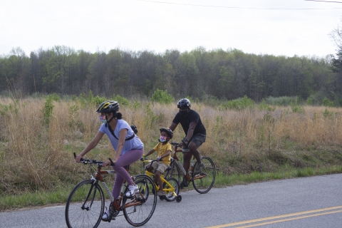 Two adults biking a paved road with a child riding with training wheels. They are next to a meadow.