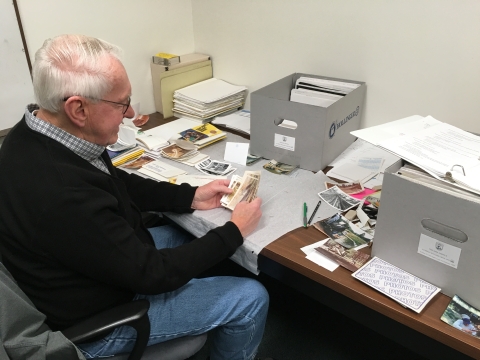A man sits at a desk holding some photographs. Spread out in front of him are archival folders, boxes and more photos that he is sorting to be processed and put into archival storage.