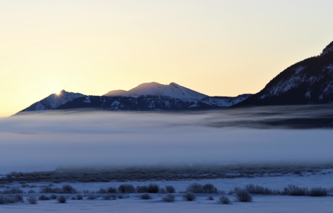 The sun peeks over the mountains during a snowy sunrise with a layer of fog blanketing the Refuge valley