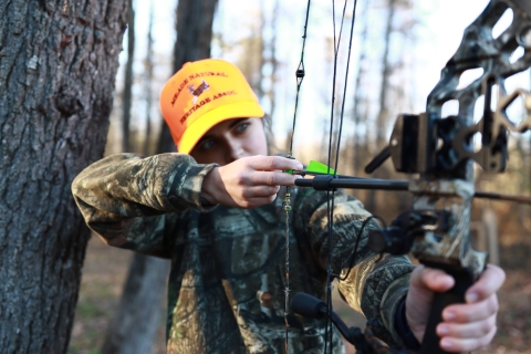 Young female about to draw bow and shoot to her left. Wearing camo and orange hat.