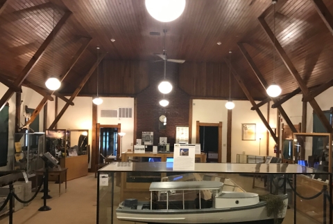 This color photo shows a view of the main exhibit room of the National Fish Culture Museum. It has a high vaulted wood ceiling with wooden support beans along the walls. Various displays can been seen with the one closet to the viewer of a large white boat.