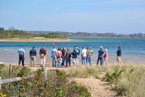 A group of about a dozen visitors stand on a beach, alongside turquoise water, and listen to an interpretive talk. 