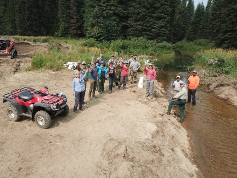 A dozen people are standing on a stream bank, and an ATV is in the left side of the frame. The people are looking at the camera.