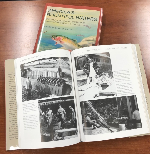 Two books are shown laying atop a table. One book is displayed open and is resting atop a closed copy of the same book, which shows the cover. The book that is open shows a photo spread of historic black and white photos of fisheries work.