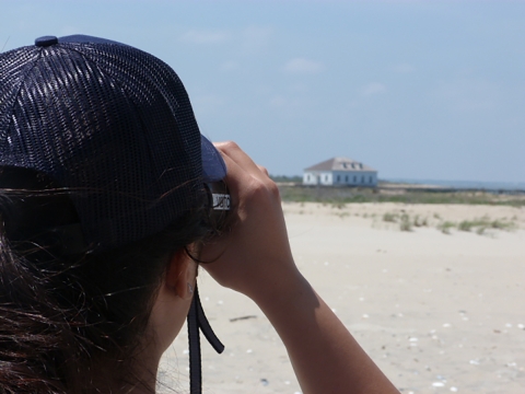 An intern uses binoculars to view a distant building on the beach