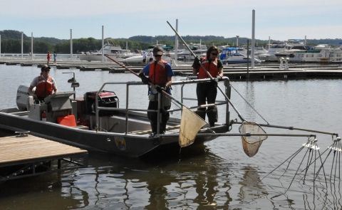 People in brown uniforms and orange flotation gear stand on a boat in a waterway holding nets.