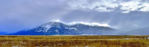 A flat, brown landscape with cloud-shrouded, snow-capped mountain in the background. Rays of sunlight are coming through the clouds.