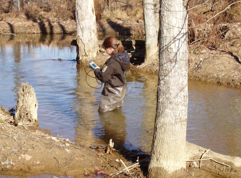 A U.S. Fish and Wildlife Service biologist wading in a narrow stream in Virginia in winter reads a handheld device that collects water quality data.