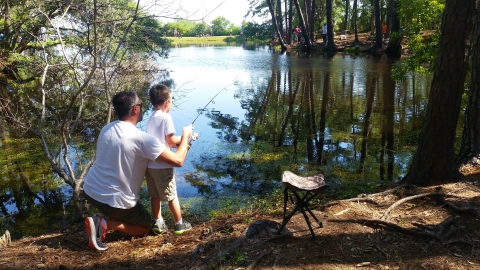 A man is kneeling next to a young boy teaching him how to hold a fishing rod while trying to fish in a shaded pond.