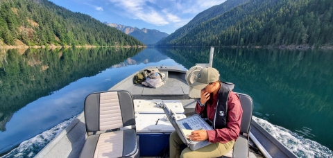 A person in waders sits in a moving motorized boat in a lake, looking at an open laptop.