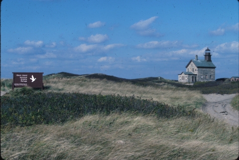 Looking out across waving grass at a lighthouse in the distance on Block Island Refuge, identified by a brown sign.
