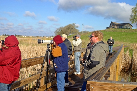 A group of people with binoculars and cameras observe distant waterfowl while standing on a wood observation deck.