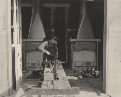 Black and white photo of a man in overalls working on pipes in front of 2 furnaces with very large, tall chimneys.