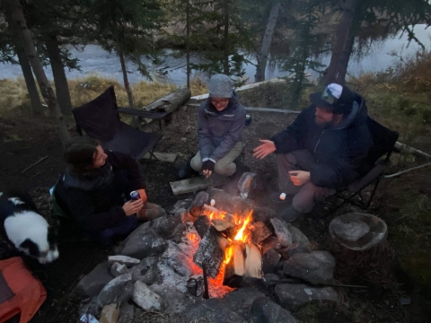 Two women, a man, and a small dog talk around a glowing campfire