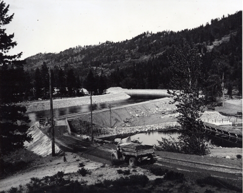 Black and white photo of a dump truck driving up onto a constructed berm with water in a newly built channel beyond.