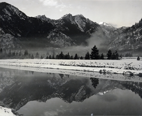 Black and white photo of mountains reflected in a constructed channel on a snowy winter day.