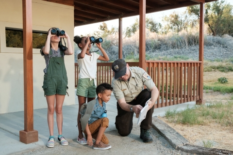 A ranger kneels down while pointing to a book. Two children look through binoculars.