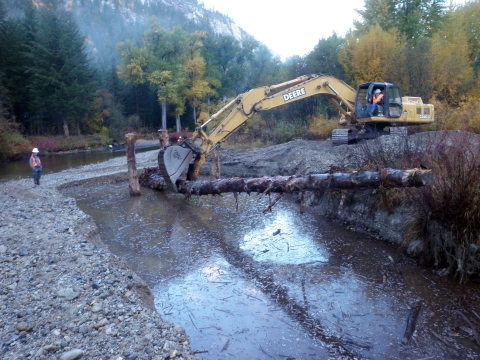 Photo of a backhoe fully extended to lay a log in a stream bed. One person wearing a hardhat oversees nearby.