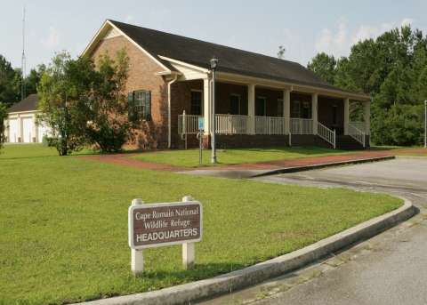 Cape Romain NWR Headquarters building with small headquarters sign in front lawn area close to driveway and parking lot.