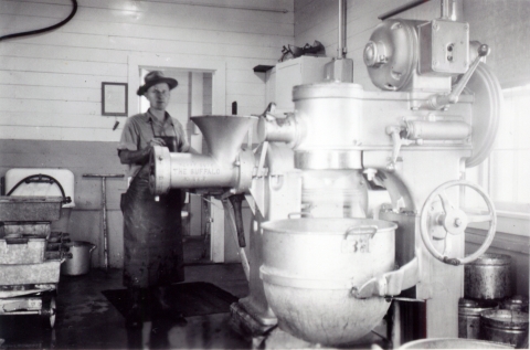 Black and white photo of a man in overalls and round-rimmed hat standing beside an enormous meat grinder.