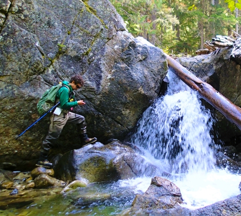 A young man in waders with a backpack stands near a waterfall.