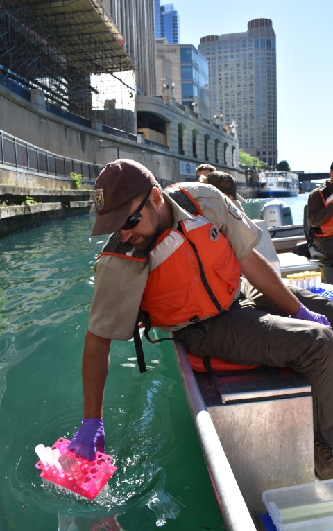 A person in an orange vest and brown uniform collects water into a vial from a boat with a city riverfront and skyline in the background