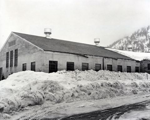 Black and white photo of a concrete building with tall windows, surrounded by large piles of snow.