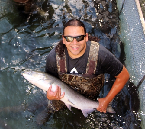 Smiling man in sunglasses and waders holds a large salmon by the tail and over one arm.