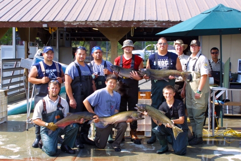 A group of 11 men of varying age stand together in waders, holding up salmon and smiling in a pose for the camera.