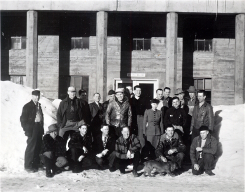 Black and white photo of a group of 21 men and 1 woman in from of a concrete building with pillars with banks of snow at least 6 feet high on either side of the entrance.