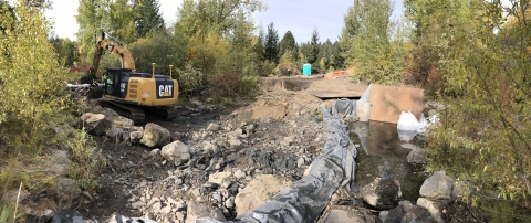 An excavator works in a stream channel blocked with boulders to hold back water.