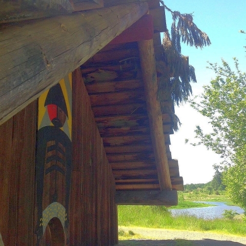 An A-frame log and plank building with Native American art above its doorway and a body of water next to it