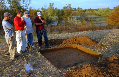 Four men looking at a small, shallow, square archaeological site next to a body of water