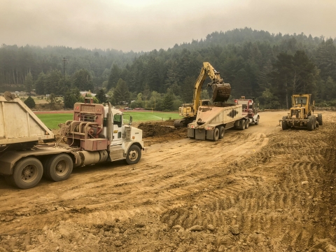 Tractor trailer with bulldozers and other construction equipment along dirt road on cloudy day with forest in background