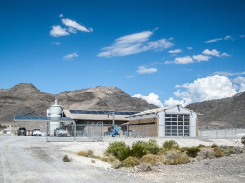 A building with solar cells on roof, three vehicles parked to the side, surrounded by a chain link fence. Desert mountains in the background and a blue cloudy sky 