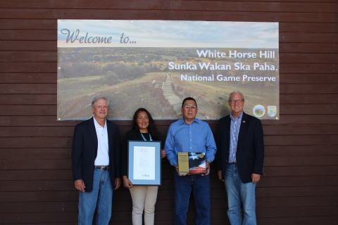 Four people stand in a line in front of a sign that reads: Welcome to White Horse Hill Sunka Wakan Ska Paha National Game Preserve