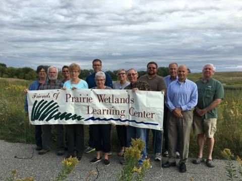 12 adults outside holding a banner with the words "Friends of the Prairie Wetlands Learning Center"