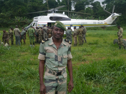 A Gabonese military officer faces the camera, standing on a field with a group of people in khaki uniforms standing near a helicopter in the background.