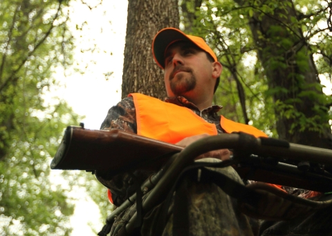 A ground-up view of a hunter in camouflage and an orange blazer sitting with a gun in his lap