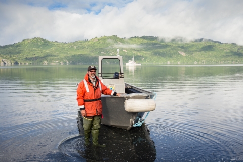 A man in an orange coat stands in the water next to an aluminum skiff.