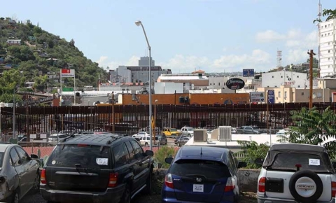 A tall border wall is shown with buildings behind it and cars parked in front of it.
