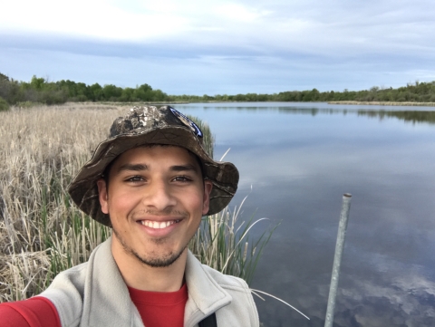 A young man in a vest and hat stands on riverbank and smiles at camera.