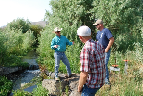 Three men stand by a creek. The man on the left has a cowboy hat on and is using hand gestures. The other two men are facing the cowboy.