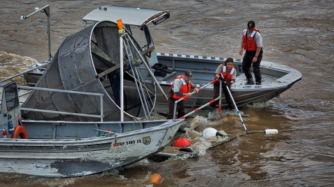 Three people on a boat in a river work to maneuver a large piece of equipment