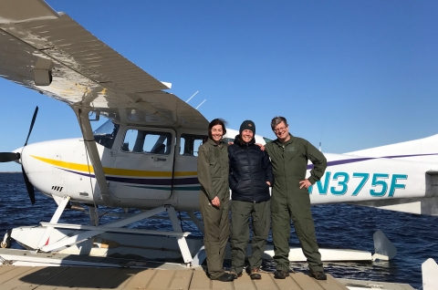 two women and one man standing in front of amphibious cessna 207 aircraft which is white with yellow, purple, blue stripes