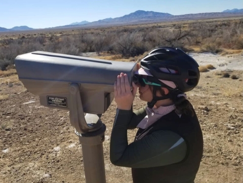 a woman wearing a cycling helmet looks through stationary binoculars across the desert