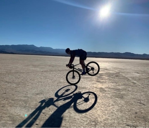 A cyclist's silhouette and shadow are captured on a dry, desert lake bed surrounded by mountains. 